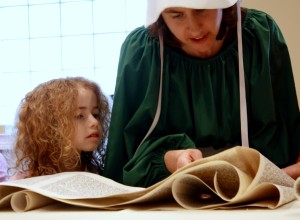 Woman in costume reading the megillah with her young daughter beside her, watching.